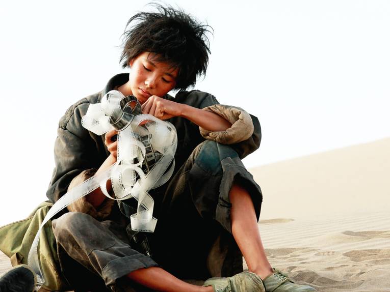 Ein junger Mensch sitzt auf einer Düne und begutachtet einen verhedderten 35mm Film, den er in den Händen hält.